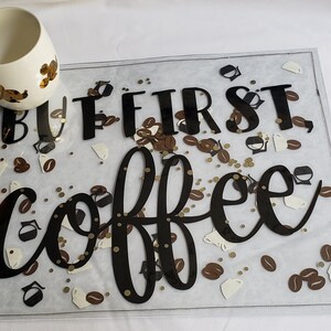 May include: A clear plastic sign with the text "But first, coffee" in black lettering. The sign is decorated with gold glitter and brown coffee bean cutouts. A white coffee mug with gold accents is in the foreground.