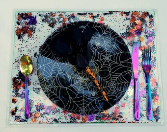 May include: A black and white spiderweb patterned dinner plate with a black witch's hat and a gold spoon, a purple fork and a purple knife on a glittery white background with orange, red, and green confetti.