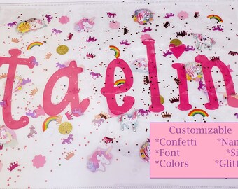 May include: A pink and white personalized name sign with the name "taelin" in pink letters. The sign is decorated with glitter, confetti, and unicorn stickers. Customizable with different confetti, font, colors, and glitter.