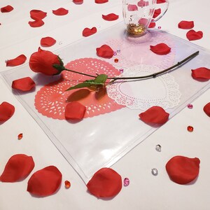 May include: A clear plastic sheet with a red rose and white doilies on top. The doilies are arranged in a circle with the rose in the center. There are red rose petals scattered around the sheet.