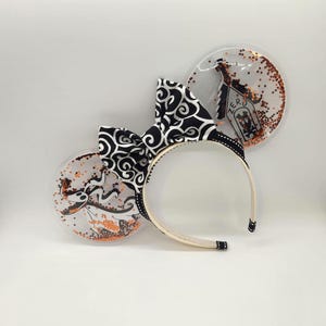 May include: Black and white patterned fabric bow headband with two clear plastic ears. One ear features a black and white image of Jack Skellington from The Nightmare Before Christmas. The other ear features a clear plastic house with the word "Zero" on it. Both ears have orange glitter inside.