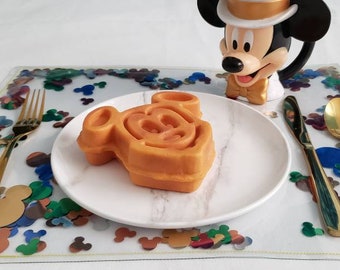 May include: A white plate with a Mickey Mouse shaped waffle on it. The plate is on a clear placemat with colorful confetti. There is a gold fork and spoon on the placemat.
