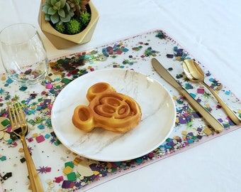 May include: A white plate with a Mickey Mouse shaped waffle on a confetti-covered placemat with gold silverware.