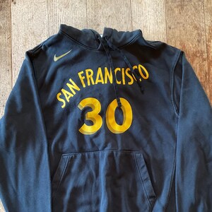 May include: Navy blue hoodie with the words "SAN FRANCISCO" and the number "30" in yellow. The Nike logo is visible on the upper left chest. The hoodie has a front pocket and a drawstring hood.