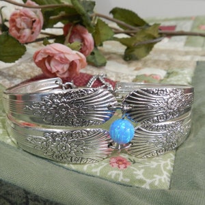 May include: A silver bracelet made from repurposed silverware, featuring a floral design. The bracelet has a blue opal-like bead as a focal point. The bracelet is displayed on a patchwork quilt with pink roses in the background.