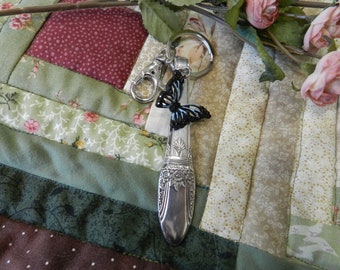 May include: A silver spoon keychain with a decorative butterfly charm. The butterfly is black with blue accents. The spoon has ornate floral detailing and a silver clasp.