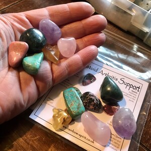 May include: A collection of various colored crystals, including pink, green, purple, and yellow, in a hand. The crystals are labeled with their names and healing properties on a white card that reads "Apatoid Arthritis Support".