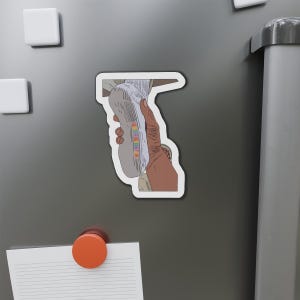 May include: A sticker featuring a hand holding a sneaker. The sneaker is light gray with rainbow text that reads "BE DEE DEE N". The sticker is on a gray surface with white square magnets and an orange circular magnet.