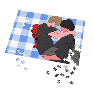 Op de afbeelding: Een rechthoekige legpuzzel met een blauw-witte vichy-ruit achtergrond en een illustratie van twee mensen die elkaar omhelzen. De een draagt een zwart shirt en een rode rugzak, de ander een zwart shirt. De tekst Austin Toney is zichtbaar.