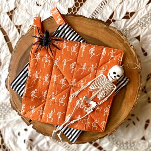 May include: Two orange and white quilted pot holders with a skeleton and spider design. The pot holders are on a wooden surface.