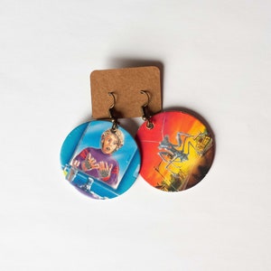 May include: A pair of round earrings with a blue and red design. One earring features a man with a shocked expression, while the other earring features a silhouette of a person standing on a platform.