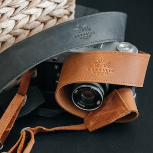 May include: A brown leather camera strap with the text "POHVALIN" embossed on it. The strap is wrapped around a silver camera with a brown leather lens cover.