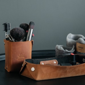 May include: A brown leather makeup brush holder with the word "POHVALIN" printed on the front. The holder is filled with various makeup brushes, including a black and silver brush, a black and white brush, and a black and brown brush. The holder is sitting on a brown leather tray with the word "POHVALIN" printed on the front.