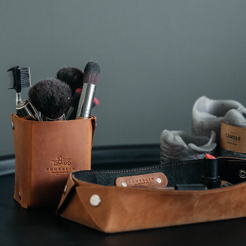 May include: A brown leather makeup brush holder with the word "POHVALIN" printed on the front. The holder is filled with various makeup brushes, including a black and silver brush, a black and white brush, and a black and brown brush. The holder is sitting on a brown leather tray with the word "POHVALIN" printed on the front.