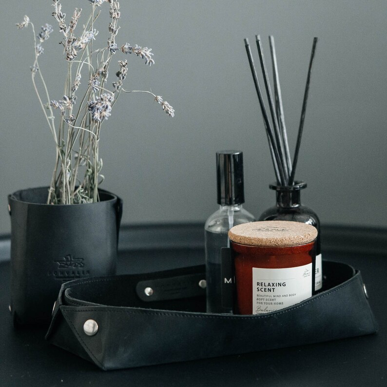 May include: A black leather tray with a black leather handle. The tray contains a brown candle with a cork lid and the text "Relaxing Scent" on the label. The candle is next to a clear glass bottle with a black cap and a black diffuser with black reeds.