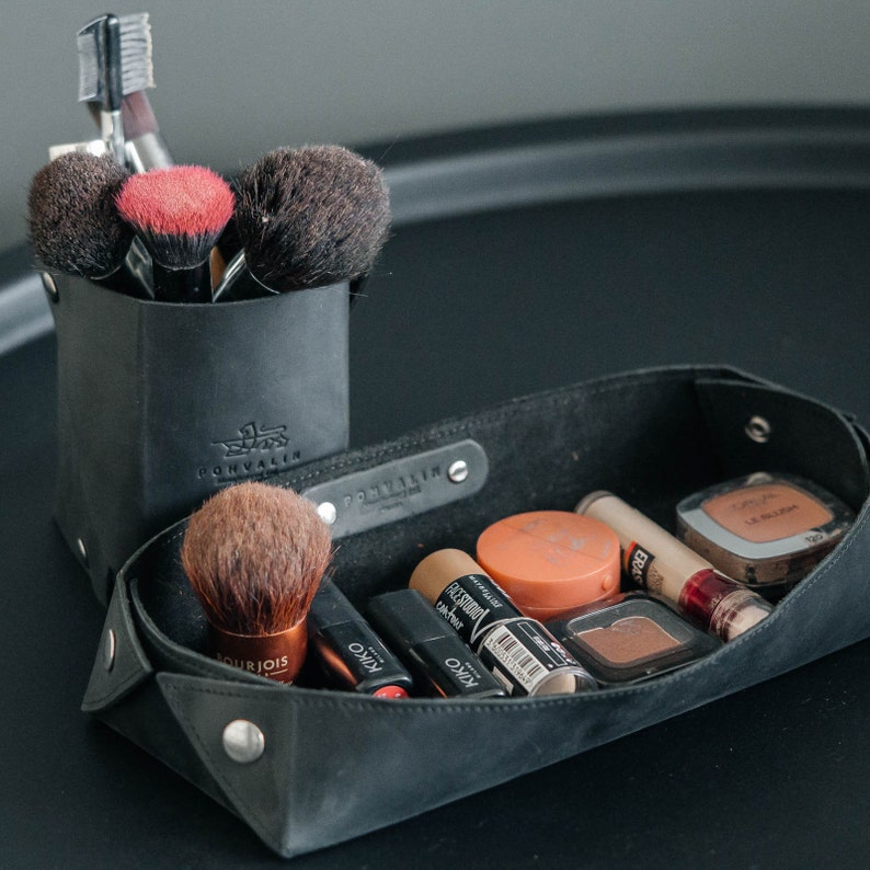 May include: A black leather makeup organizer with a tray and a separate compartment for brushes. The tray contains various makeup products, including foundation, blush, and eyeshadow.