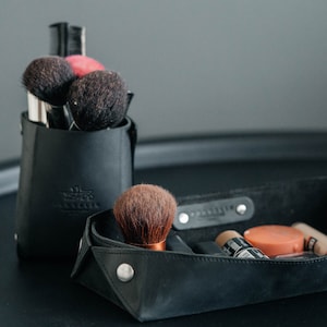 May include: A black leather makeup organizer with a variety of makeup brushes and a small tray for cosmetics. The organizer has a label that says "Traveler".