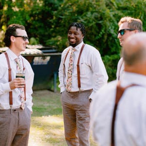 May include: A group of men in white shirts, brown braces, and floral ties, standing outdoors. They are wearing gray trousers and sunglasses. The background features green foliage. The scene suggests a relaxed, outdoor gathering.