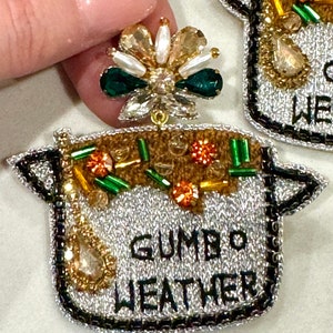 May include: Silver pot-shaped earrings with a beaded design and the text "Gumbo Weather". The earrings have a green and white beaded flower charm.