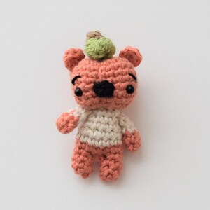 May include: A handcrafted crochet bear in peach, white, and black. The bear has a small green pom-pom on its head and is wearing a white sweater. The bear is a charming, whimsical design.