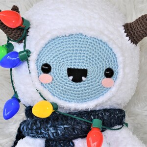 Puede incluir: Un mu&ntilde;eco de nieve de crochet blanco con cuernos de crochet marr&oacute;n, que lleva una bufanda de crochet azul y una cadena de luces navide&ntilde;as de colores.