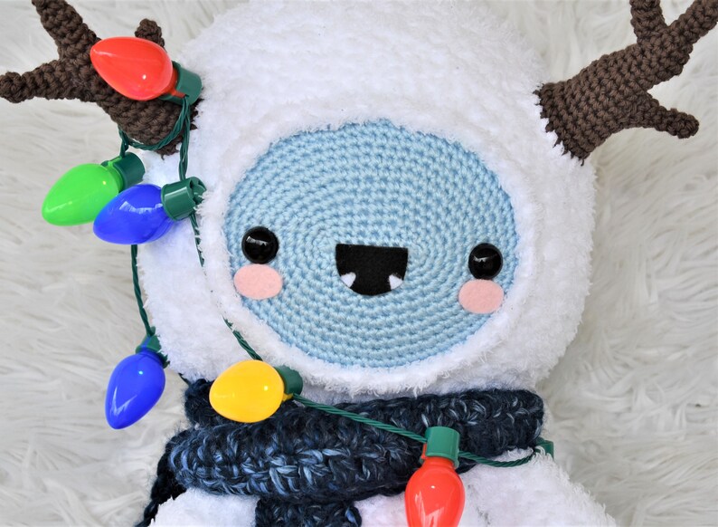 Puede incluir: Un mu&ntilde;eco de nieve de crochet blanco con cuernos de crochet marr&oacute;n, que lleva una bufanda de crochet azul y una cadena de luces navide&ntilde;as de colores.