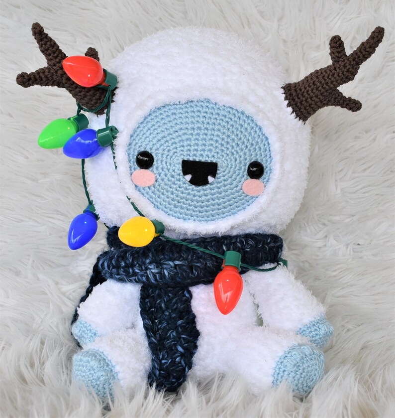 Puede incluir: Un mu&ntilde;eco de nieve de crochet blanco con cuernos marrones, que lleva una bufanda azul y sostiene una cadena de luces navide&ntilde;as de colores.