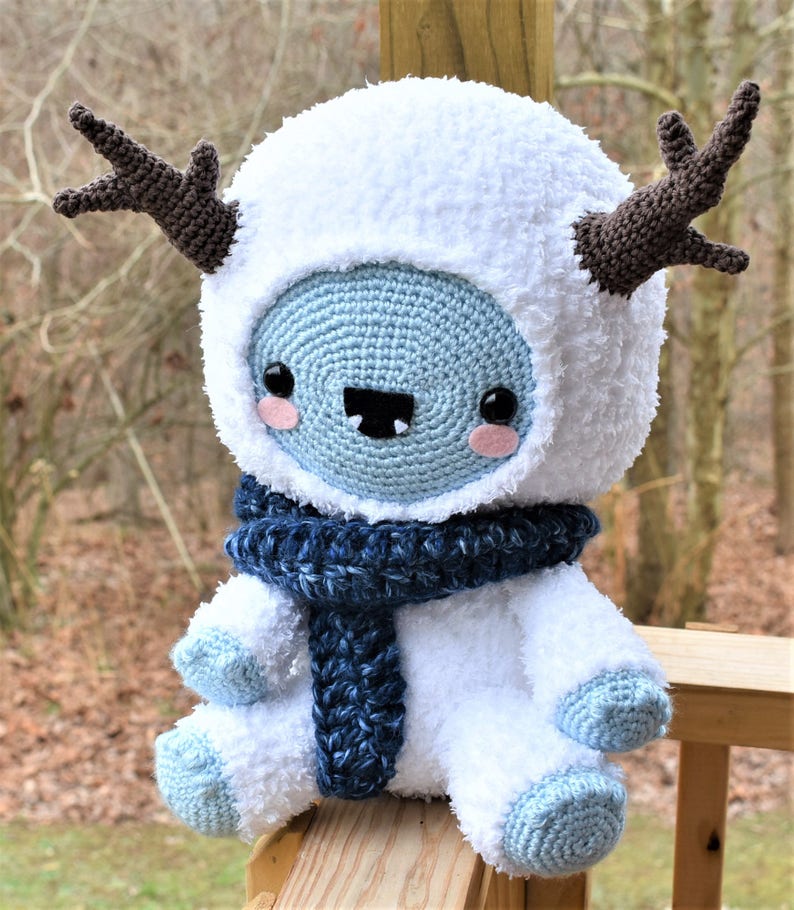 Puede incluir: Un mu&ntilde;eco de nieve de crochet blanco con detalles azules, que lleva una bufanda azul y blanca y cuernos marrones. El mu&ntilde;eco de nieve tiene una cara sonriente con mejillas rosadas y una sonrisa de dientes.