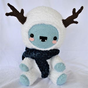Puede incluir: Un mu&ntilde;eco de nieve de crochet blanco con cuernos marrones, que lleva una bufanda azul. El mu&ntilde;eco de nieve tiene una cara azul con ojos negros, rubor rosado y una sonrisa de dientes.