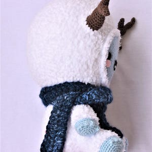 Puede incluir: Juguete de peluche blanco de crochet con cuernos marrones y una bufanda azul. El juguete tiene una cara azul claro con mejillas rosadas.