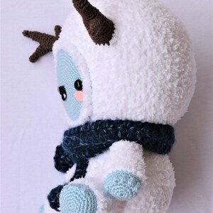 Puede incluir: Un juguete de peluche blanco tejido a crochet con cuernos marrones, una bufanda azul y pies azul claro. El juguete tiene una cara linda de estilo de dibujos animados con ojos negros y mejillas rosadas.