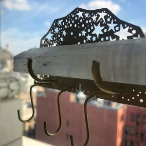 Op de afbeelding: Een gouden metalen wandbevestigde sleutelhanger met een stervormig ontwerp en drie haken. De sleutelhanger is gemonteerd op een wit selenietkristal plank.
