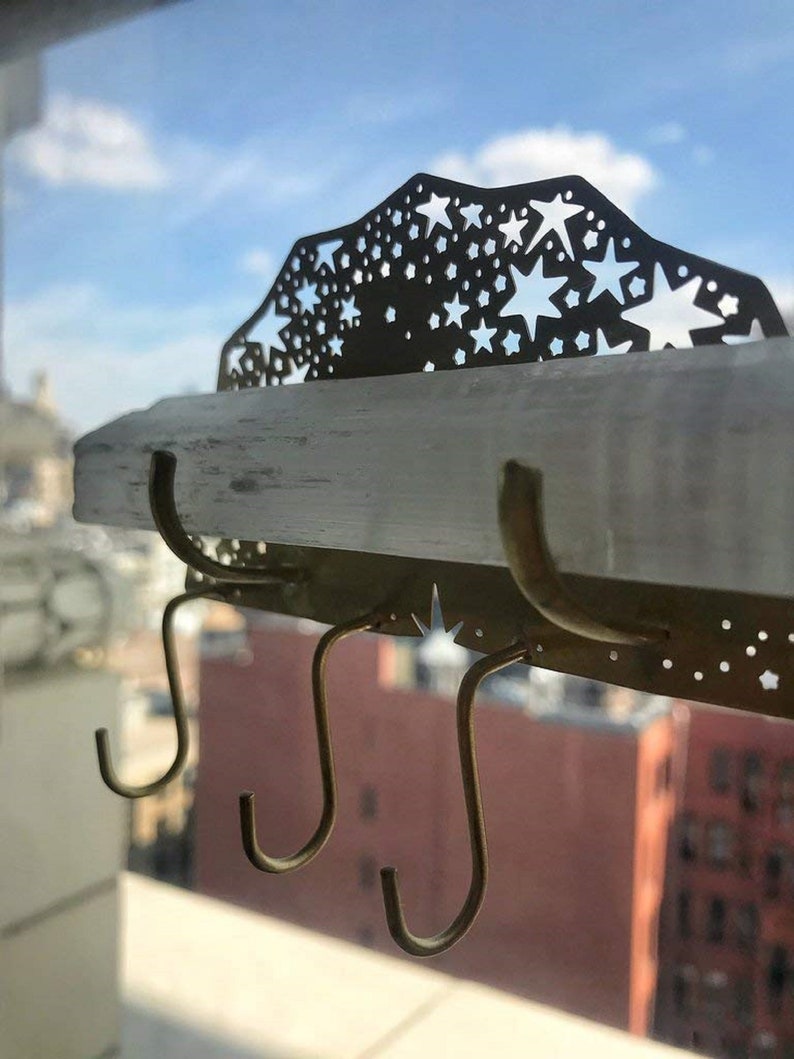 Op de afbeelding: Een gouden metalen wandbevestigde sleutelhanger met een stervormig ontwerp en drie haken. De sleutelhanger is gemonteerd op een wit selenietkristal plank.