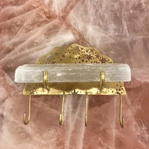 Op de afbeelding: Een gouden metalen wandbevestigde sleutelhanger met een stervormig ontwerp en een selenietkristalplank. De sleutelhanger heeft vier haken.
