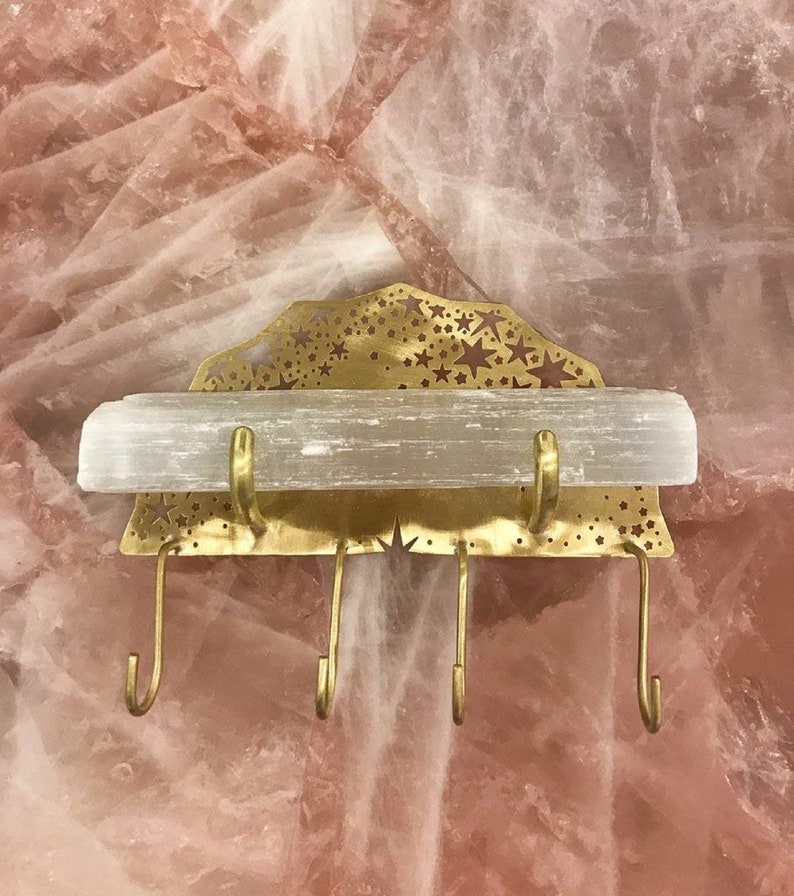 Op de afbeelding: Een gouden metalen wandbevestigde sleutelhanger met een stervormig ontwerp en een selenietkristalplank. De sleutelhanger heeft vier haken.