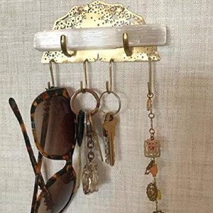 Op de afbeelding: Een messing sleutelhanger met een decoratief ontwerp en een wit kristallen plankje. De hanger heeft vijf haken en hangt aan een muur. Een paar zonnebrillen, sleutels en een armband met bedels hangen aan de haken.