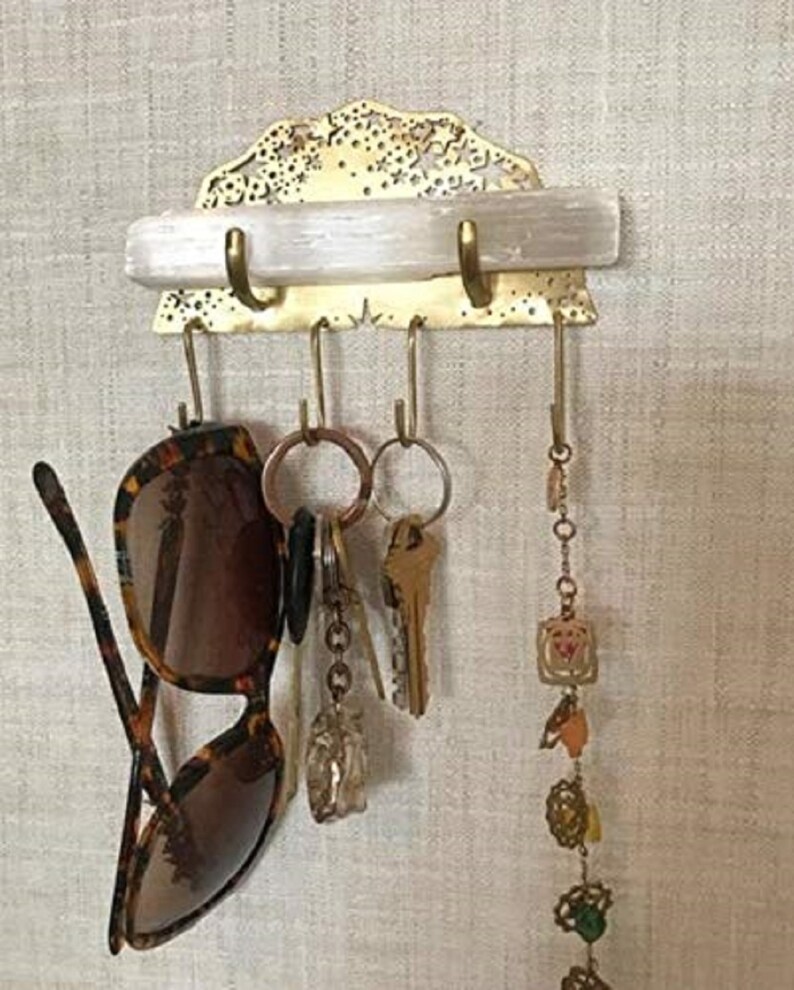 Op de afbeelding: Een messing sleutelhanger met een decoratief ontwerp en een wit kristallen plankje. De hanger heeft vijf haken en hangt aan een muur. Een paar zonnebrillen, sleutels en een armband met bedels hangen aan de haken.