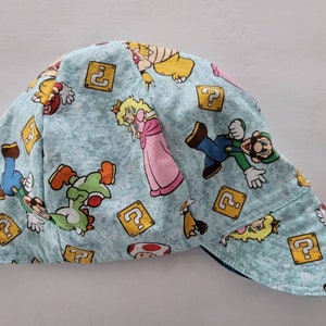 Könnte beinhalten: Hellblaue Schweißerkappe mit einem sich wiederholenden Muster von Charakteren aus Super Mario Bros., darunter Mario, Luigi, Prinzessin Peach und Yoshi, sowie Fragezeichenblöcken.