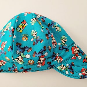 May include: A teal welding cap featuring a repeating pattern of Mario and Luigi characters, along with the words "Super Mario". The cap has a curved brim and is stitched with black thread.