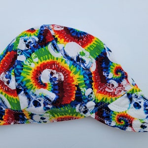 Può includere: Un cappuccio da saldatore colorato con un motivo tie-dye con teschi bianchi. Il cappuccio ha una tesa curva ed è realizzato in tessuto con una cucitura. Il design include vortici di rosso, giallo, verde e blu.