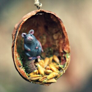 May include: A whimsical miniature mouse figurine inside a walnut shell, suspended by a twine string. The mouse is gray with pink ears and paws, nestled among seeds and moss. The shell is brown with a textured surface, creating a rustic, natural aesthetic.