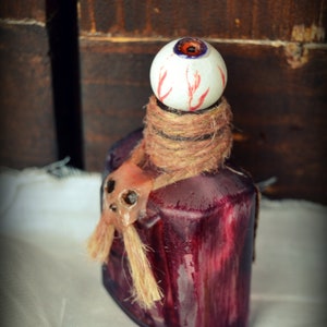 May include: A dark red, square-shaped bottle with a white eyeball stopper, featuring red veins. The bottle is wrapped with twine and has a small, decorative skull attached. The background is a dark wooden surface.
