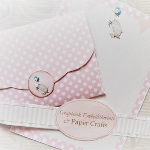 Puede incluir: Un sobre rosa con lunares blancos y un borde festoneado. El sobre tiene una pegatina circular con un pato de dibujos animados. El sobre está al lado de una tarjeta blanca con un borde rosa y un pato de dibujos animados. La tarjeta tiene el texto "Scrapbook Embellishments & Paper Crafts".