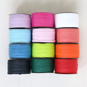 Può includere: Una collezione di rocchetti colorati di nastro di rafia in varie tonalità, tra cui lavanda, rosa, bianco, azzurro, corallo, blu, verde, arancione, nero, verde smeraldo e rosso. I rocchetti sono impilati, mostrando la gamma di colori.