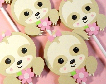 May include: Six brown sloth cupcake toppers with pink bows and flowers. The sloths have black eyes and a green collar.