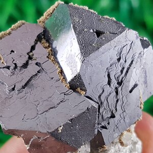 Puede incluir: Un grupo de cristales octa&eacute;dricos met&aacute;licos de color gris oscuro con un cristal verde en el centro. Los cristales tienen una superficie rugosa y texturizada.