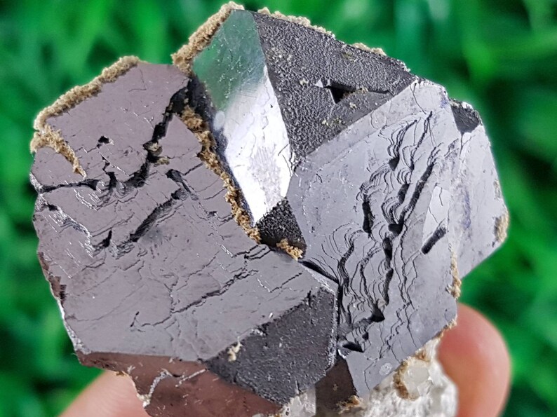 Puede incluir: Un grupo de cristales octa&eacute;dricos met&aacute;licos de color gris oscuro con un cristal verde en el centro. Los cristales tienen una superficie rugosa y texturizada.