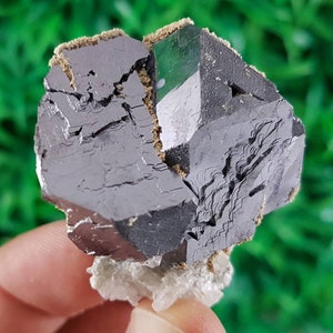 Puede incluir: Un gran cristal met&aacute;lico gris oscuro con forma geom&eacute;trica. El cristal tiene una superficie rugosa y texturizada y parece estar parcialmente roto. El cristal est&aacute; sentado sobre una base cristalina blanca.