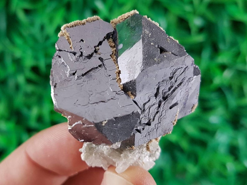 Puede incluir: Un gran cristal met&aacute;lico gris oscuro con forma geom&eacute;trica. El cristal tiene una superficie rugosa y texturizada y parece estar parcialmente roto. El cristal est&aacute; sentado sobre una base cristalina blanca.