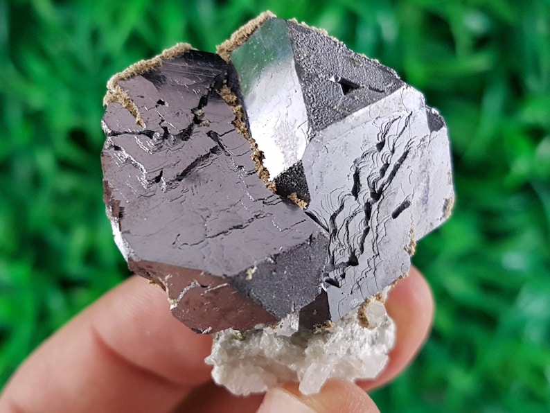 Puede incluir: Un cristal met&aacute;lico gris oscuro con forma geom&eacute;trica. El cristal tiene una superficie rugosa y texturizada y est&aacute; colocado sobre una base blanca y cristalina.